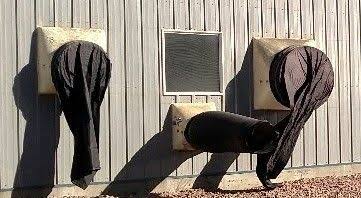 Three fans on a barn sidewall: a small minimum ventilation fan running with its air chute inflated, and two larger idle fans with their chutes pulled flat against the openings